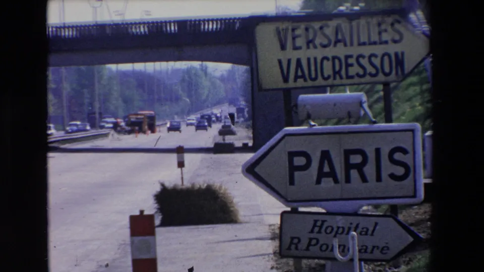 Tight 1969 Paris-region frame showing large Paris arrow signage below a Versailles and Vaucresson sign, with highway traffic and an overpass behind.