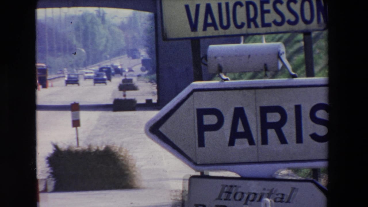 1969 Paris-area road scene with a large arrow sign pointing to Paris, a Vaucresson sign above, and distant vehicles on a tree-lined roadway.