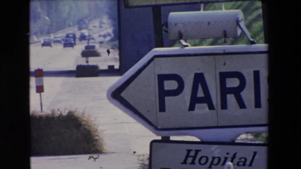 Soft, grainy 1969 Paris-area road frame showing a cropped Paris directional sign, distant traffic, and a partial hospital sign below.