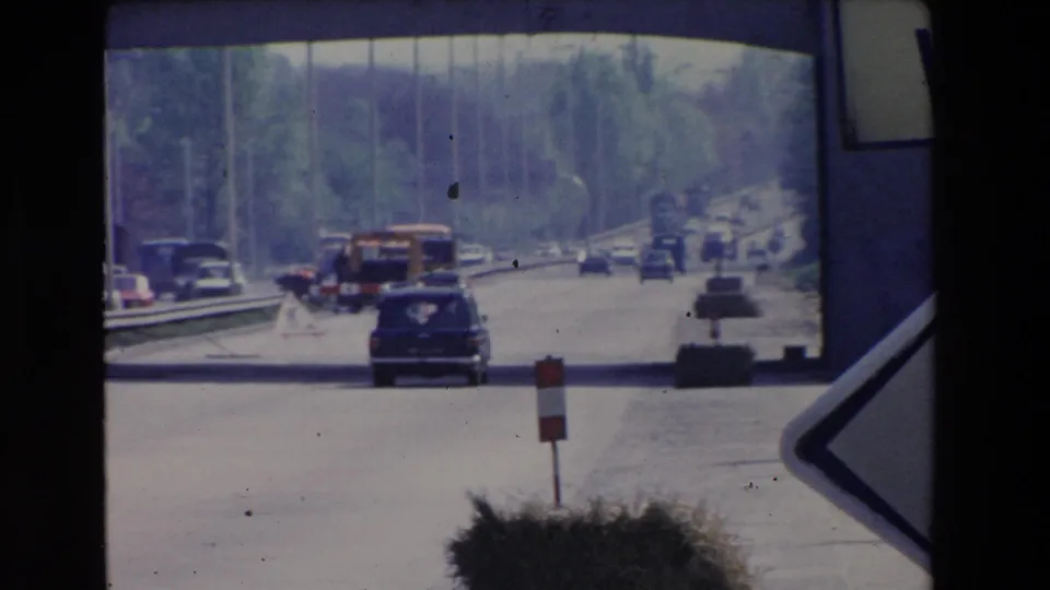 Paris-area roadway traffic in 1969 with cars and a truck moving along a tree-lined road, framed by the edge of a directional sign.