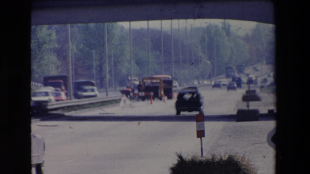 Paris, France 1969 home-movie frame showing traffic on a multi-lane roadway beneath a concrete overpass, with classic cars, trucks, trees, and road construction markers visible ahead.
