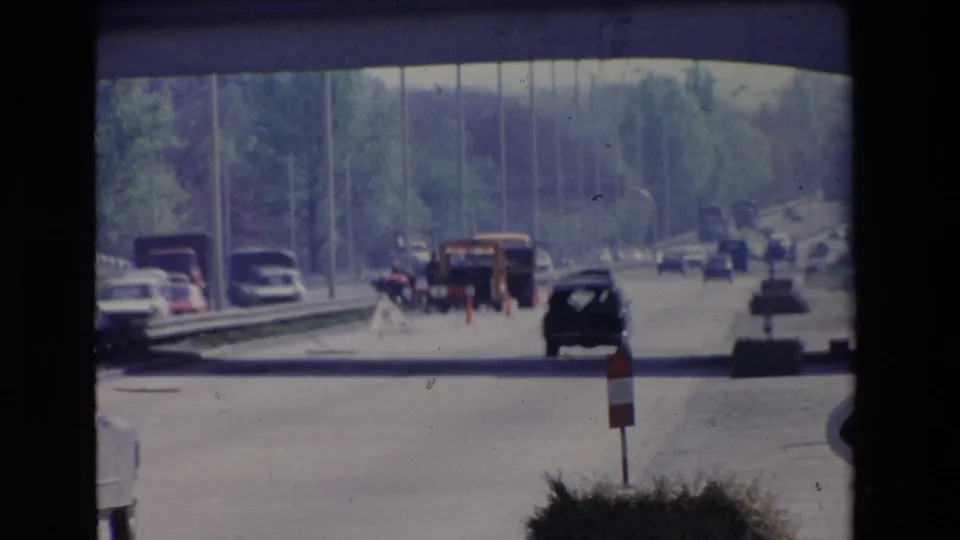 Paris, France 1969 home-movie frame showing traffic on a multi-lane roadway beneath a concrete overpass, with classic cars, trucks, trees, and road construction markers visible ahead.