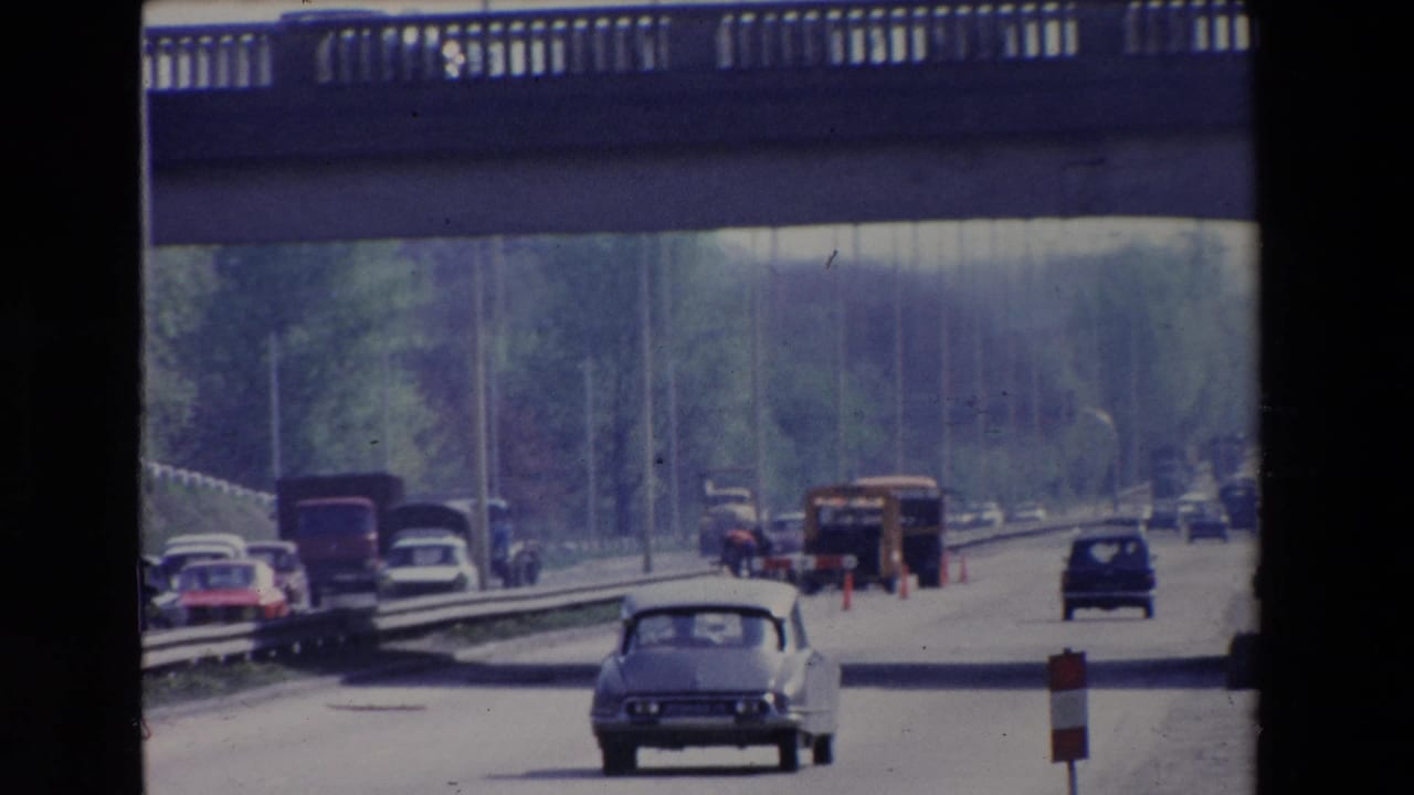 Paris, France 1969 frame with a light-colored sedan in the foreground, other period vehicles, a concrete overpass, construction cones, and distant workers or pedestrians on the roadway.