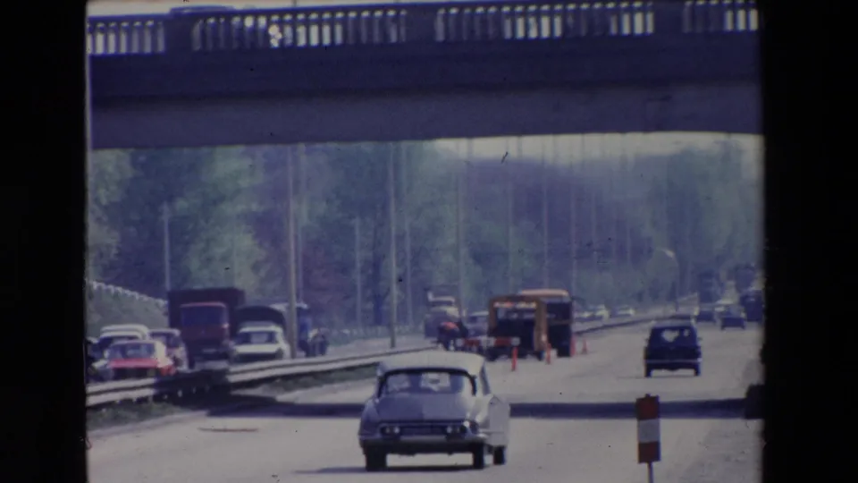 Paris, France 1969 frame with a light-colored sedan in the foreground, other period vehicles, a concrete overpass, construction cones, and distant workers or pedestrians on the roadway.