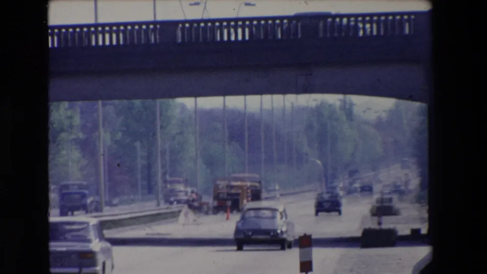 Paris, France 1969 home-movie frame showing classic automobiles on a divided multi-lane road, with a construction truck, cones, guardrail, trees, and an overpass edge in view.