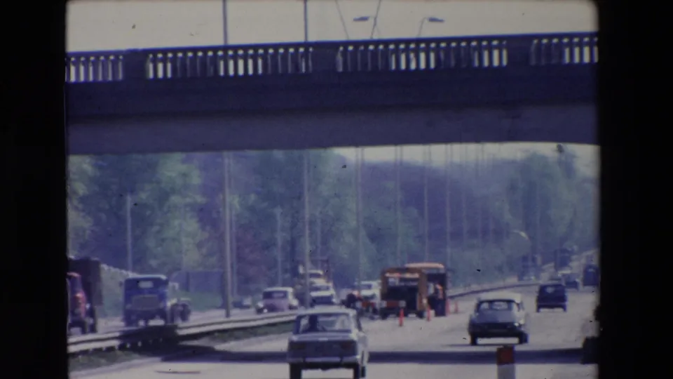 Paris, France 1969 frame of a moving-car view down a multi-lane roadway, with a sedan ahead, trucks, roadside trees, cones, temporary barriers, and a concrete overpass above.