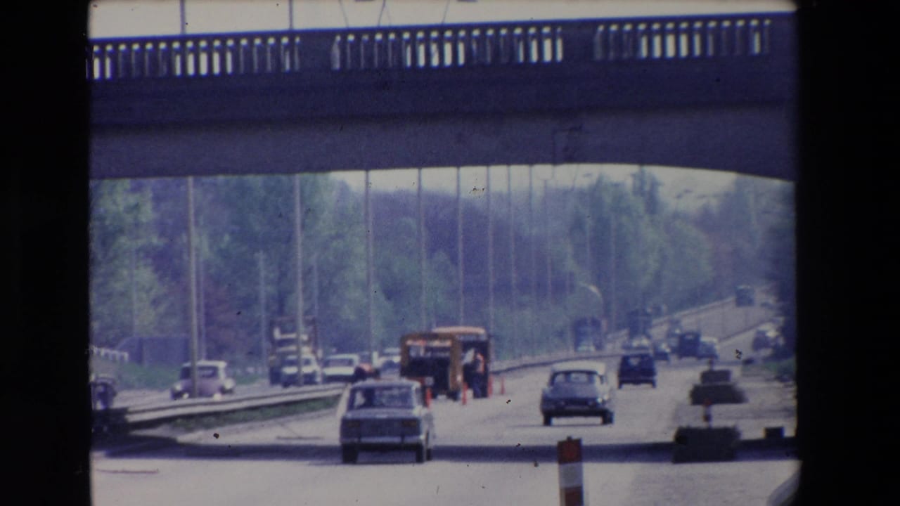 1969 Paris home-movie frame showing cars moving under a concrete overpass on a tree-lined roadway with work-zone markers ahead.