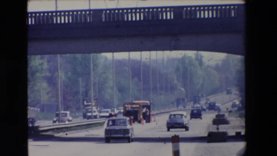 1969 Paris home-movie frame showing cars moving under a concrete overpass on a tree-lined roadway with work-zone markers ahead.