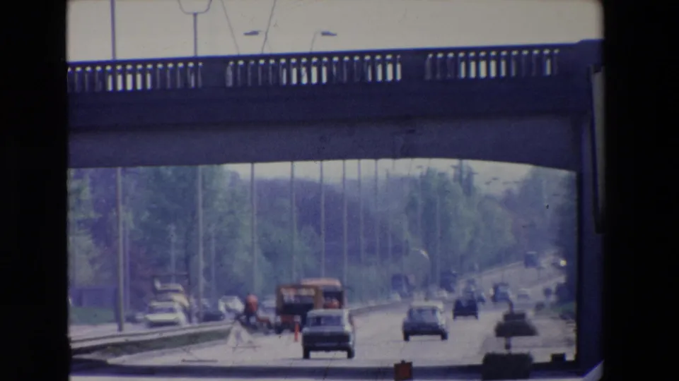 1969 Paris roadway view with cars, a bus or work vehicle, orange roadwork markers, and a concrete bridge overhead.