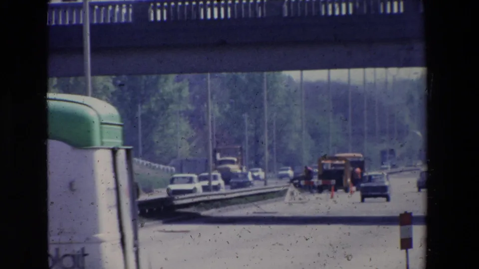 1969 Paris traffic scene with a large vehicle partly blocking the left edge, cars ahead, roadwork markers, and trees beyond the overpass.