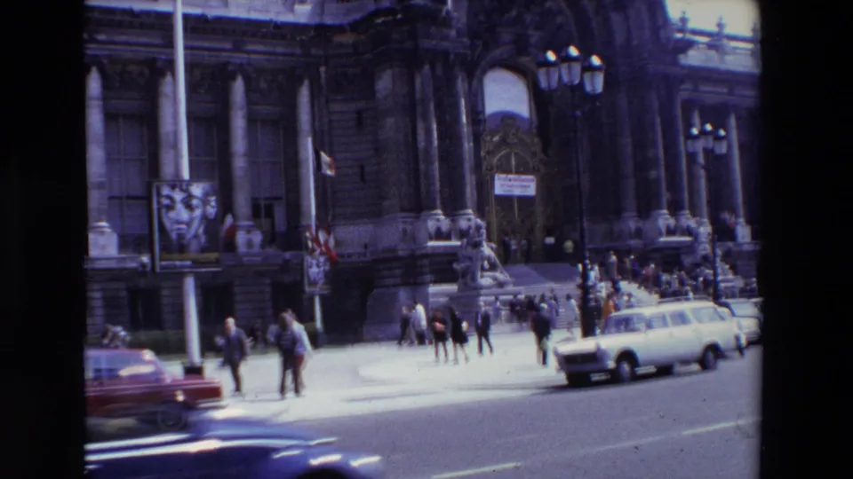 1969 Paris street view with pedestrians, cars, lampposts, flags, and an ornate public building facade visible from a moving-camera perspective.