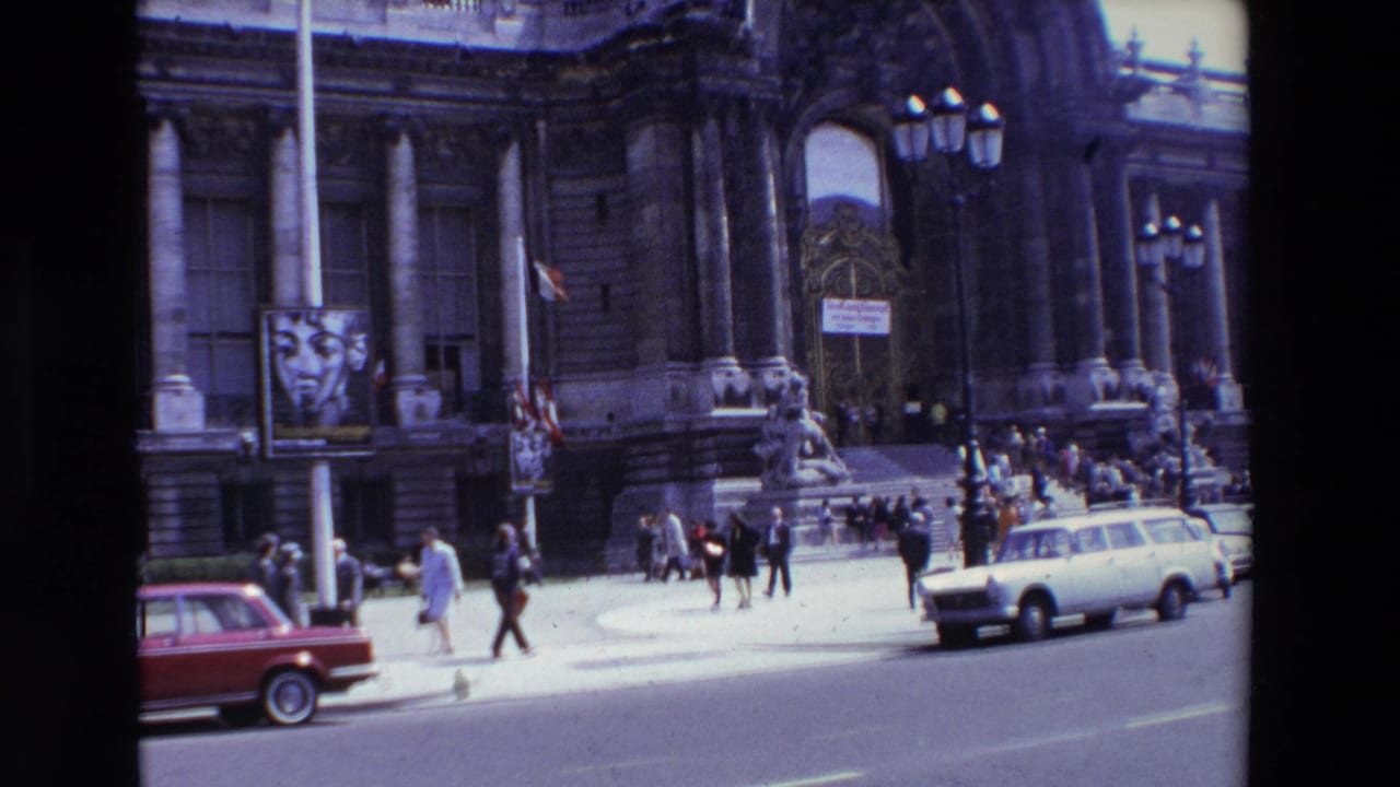 1969 Paris home-movie frame showing an ornate arched building entrance, pedestrians on the sidewalk and steps, a street poster, and passing cars.