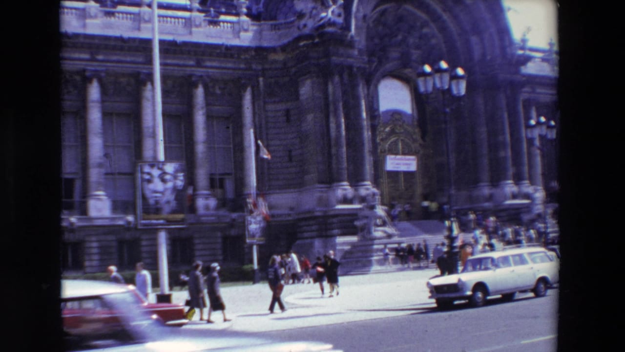 1969 Paris street view with a grand stone facade, arched entrance, pedestrians moving near the steps, and a white station wagon in bright daylight.