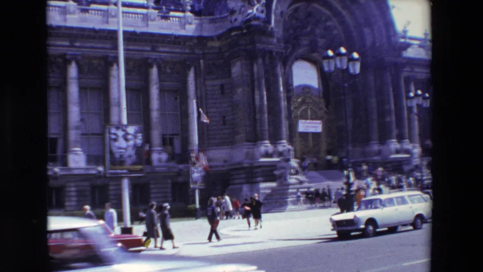 1969 Paris street view with a grand stone facade, arched entrance, pedestrians moving near the steps, and a white station wagon in bright daylight.