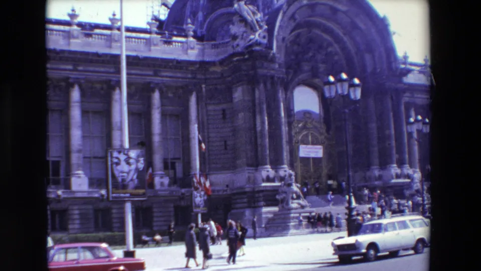 1969 Paris 8mm frame with an angled view of a massive arched portico, stone columns, pedestrians near the steps, a red sedan, and a pole-mounted poster.