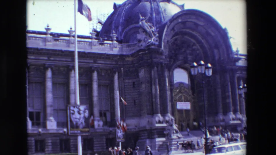 1969 Paris home-movie frame showing the upper curve of an ornate arched entrance, streetlamps, pedestrians on the steps, and part of a car at the curb.