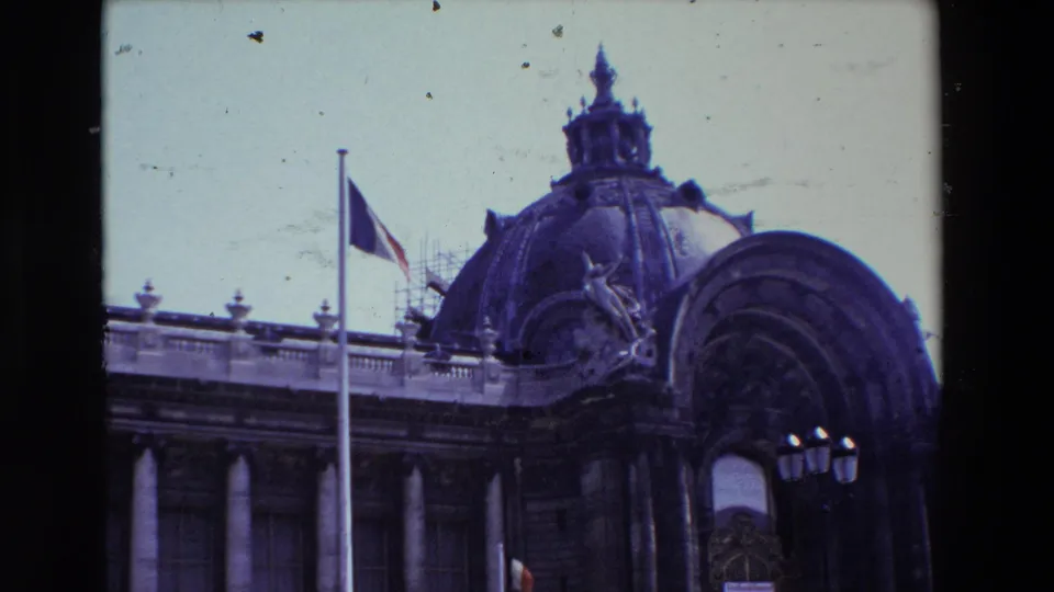 1969 Paris 8mm frame with the French flag at half-mast beside a domed public building, visible scaffolding, classical columns, rooftop sculpture, and the arched entry partly framed at right.