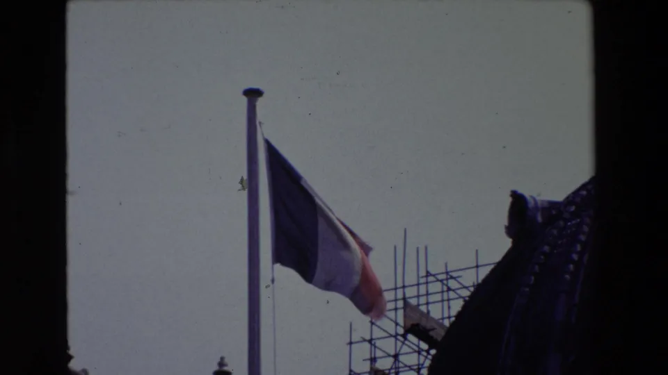 The French flag remains in view against a pale sky, with scaffolding and shadowed ornate architecture at the right edge.
