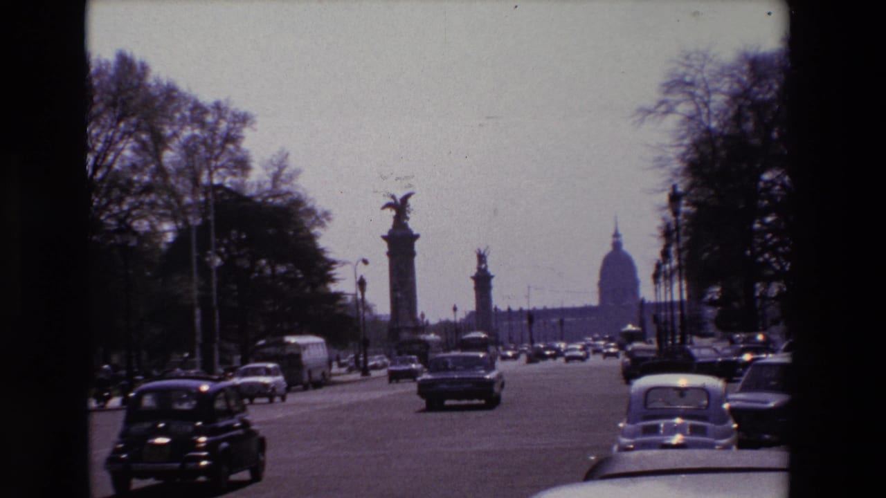 1969 Paris home-movie frame showing classic cars on a broad boulevard with statue-topped columns and the Invalides dome visible in the distance.