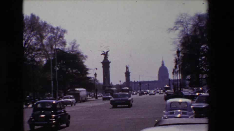 1969 Paris home-movie frame showing classic cars on a broad boulevard with statue-topped columns and the Invalides dome visible in the distance.