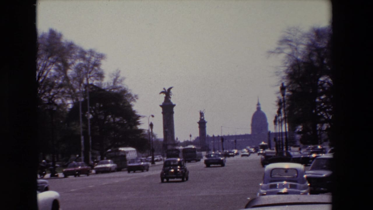 1969 Paris street view from a moving vehicle, with cars, a bus or van, ornamental columns, trees, and the Invalides dome in the background.