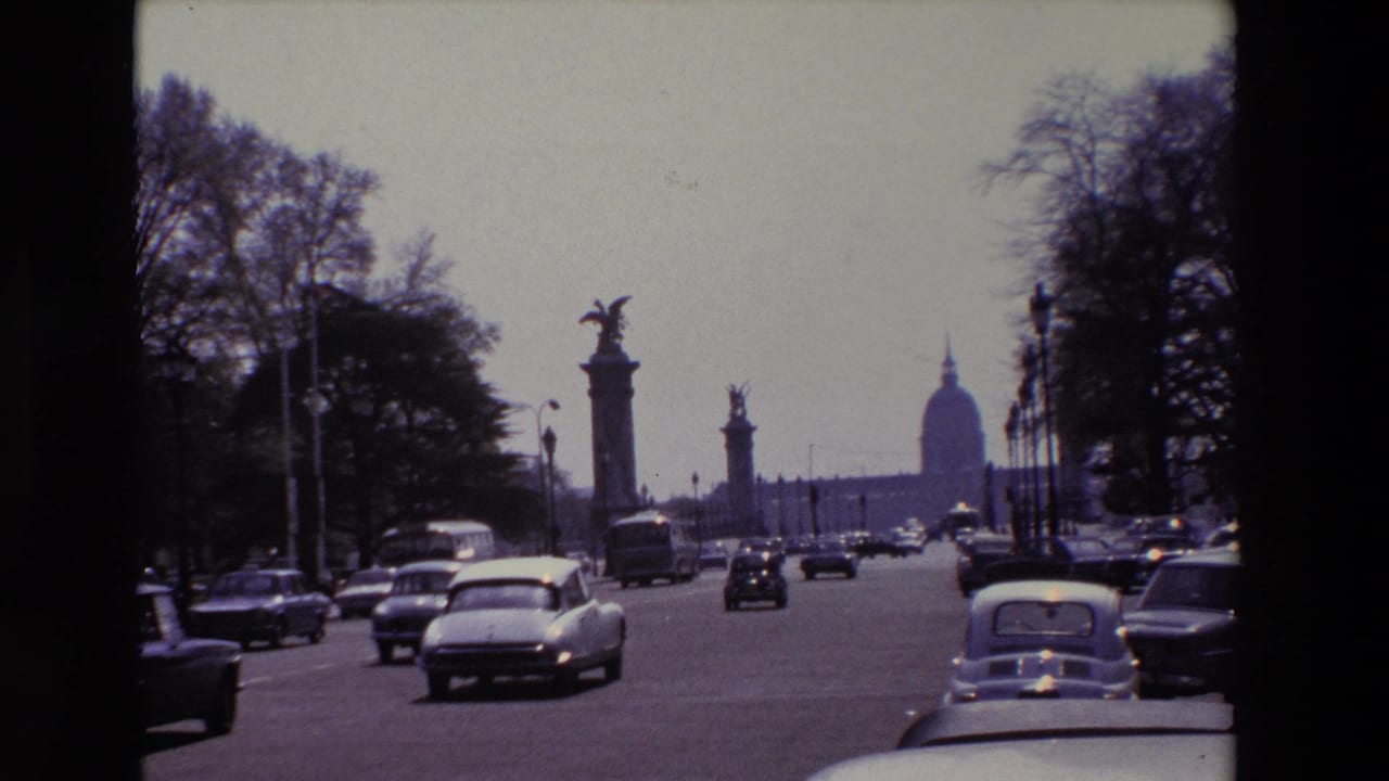 A white car and other vehicles travel on a boulevard lined with trees and monuments.