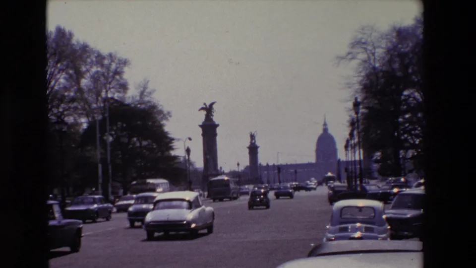 1969 Paris frame with a white car prominent in traffic, tree-lined edges, statue columns, and the Invalides dome seen ahead through haze.