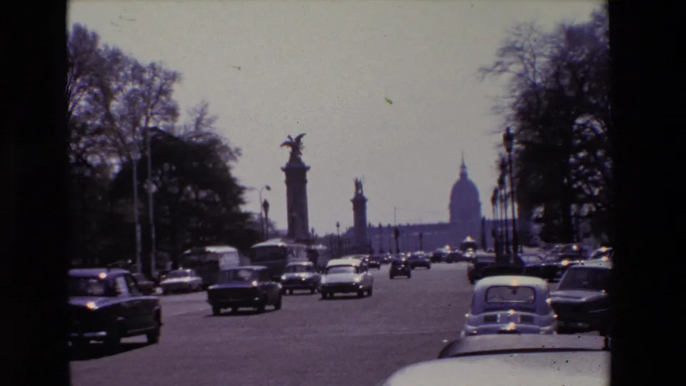 1969 Paris home-movie frame showing dense boulevard traffic, a light-colored car at the foreground edge, statue-topped columns, trees, and the Invalides dome beyond.