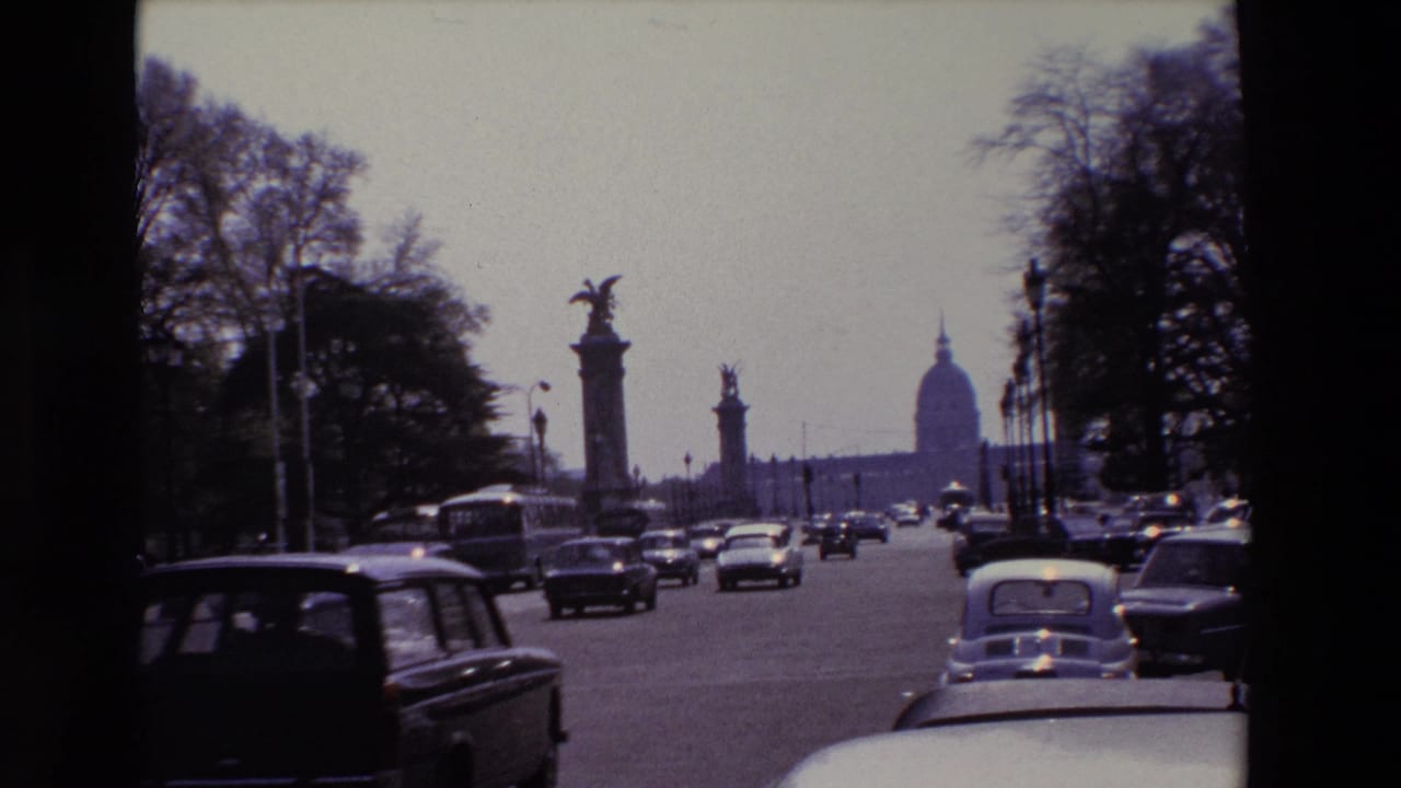 1969 Paris 8mm frame showing mid-century cars traveling along a broad avenue near stone bridge columns, with the Invalides dome visible in the distance.