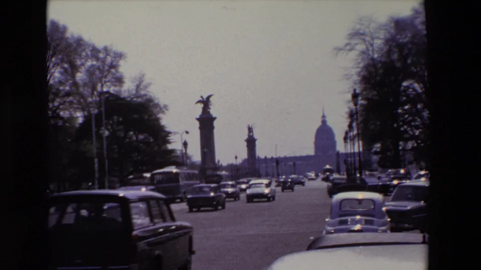 1969 Paris 8mm frame showing mid-century cars traveling along a broad avenue near stone bridge columns, with the Invalides dome visible in the distance.
