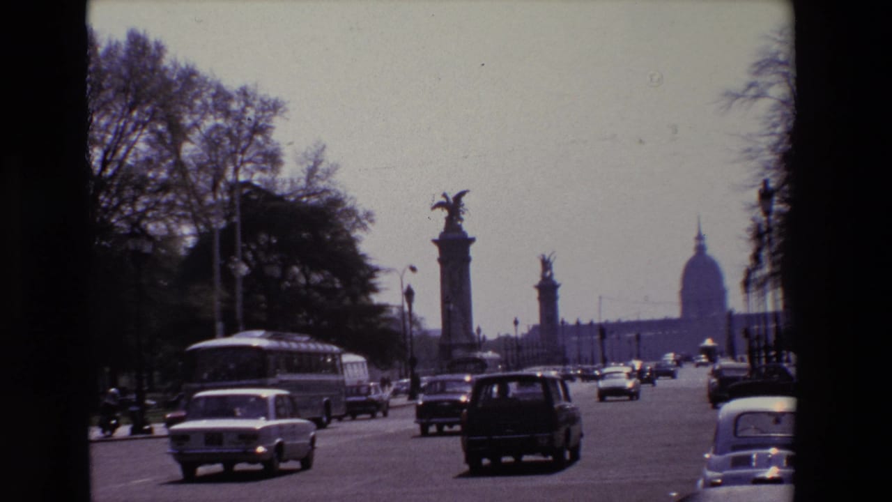 1969 Paris street view with cars and a public bus moving past tree-lined edges and tall sculptural bridge columns, with the distant Invalides dome in view.