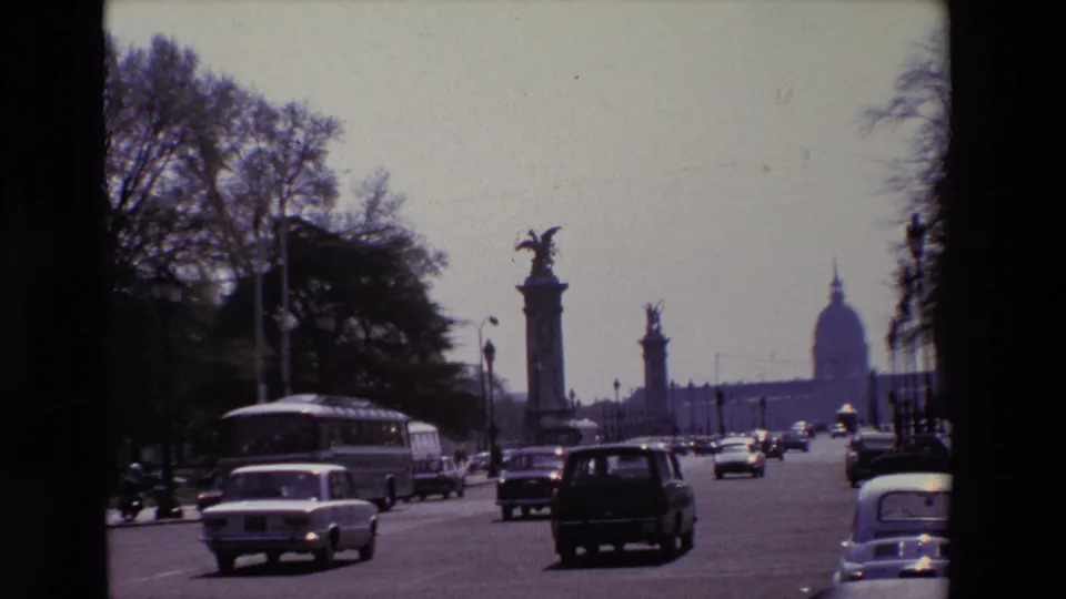 1969 Paris street view with cars and a public bus moving past tree-lined edges and tall sculptural bridge columns, with the distant Invalides dome in view.