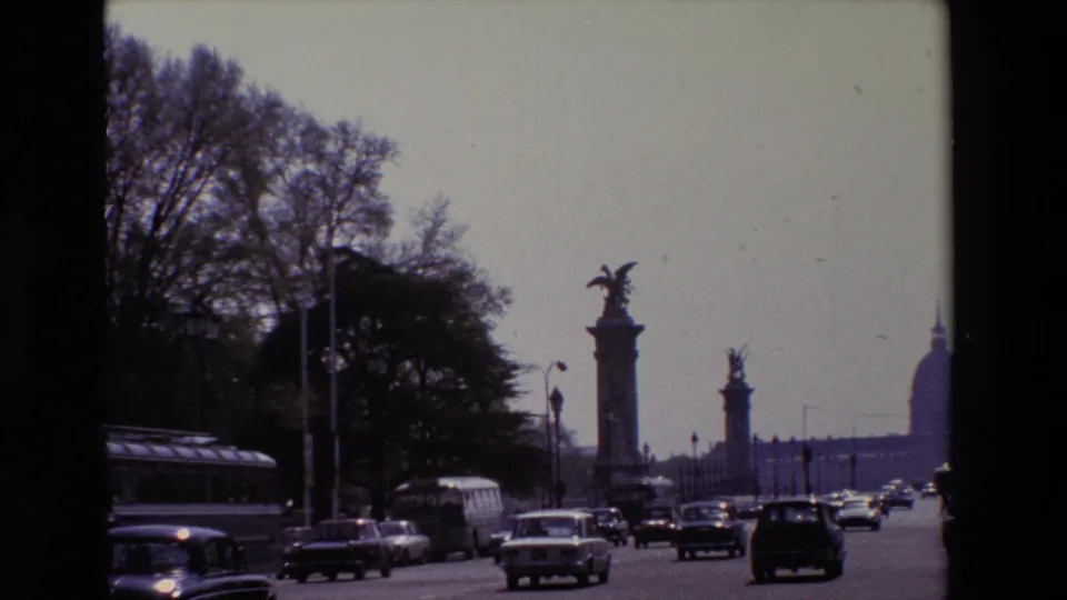 1969 Paris 8mm frame with a bus, cars, tree-lined roadside, stone columns, and a partial distant dome visible beyond the traffic corridor.