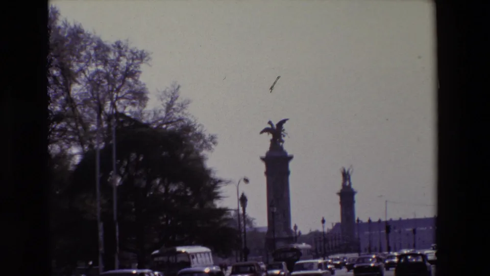 Late-1960s Paris home-movie frame showing cars, a bus, bare trees, and tall sculptural columns along the road, with soft grain and faded color.