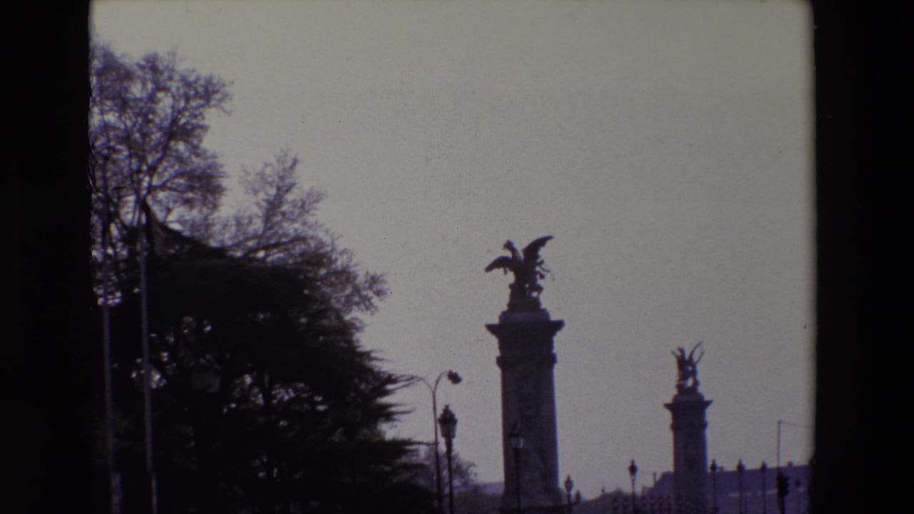 1969 Paris home-movie frame showing a silhouetted winged statue atop a tall pedestal, a second monument in the distance, dark trees, and a pale sky.