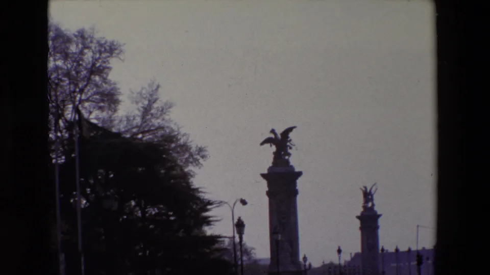1969 Paris home-movie frame showing a silhouetted winged statue atop a tall pedestal, a second monument in the distance, dark trees, and a pale sky.