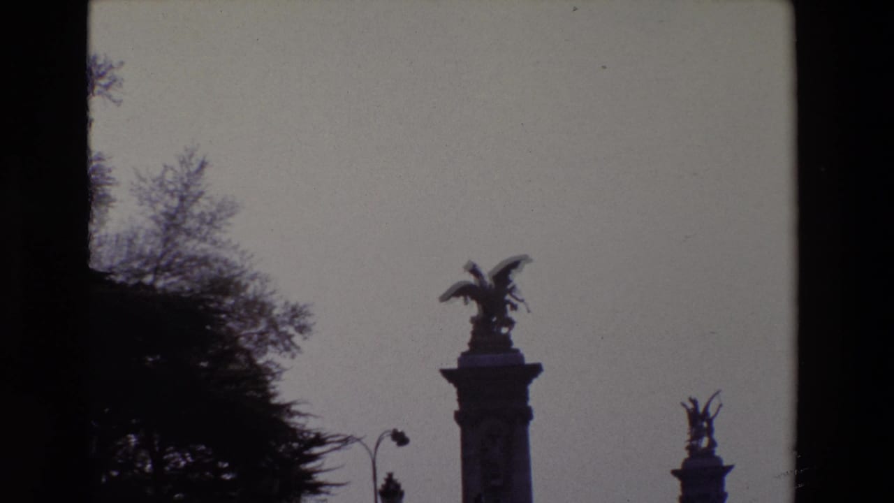 Silhouetted winged statue on a pedestal with trees and a second statue in the distance.