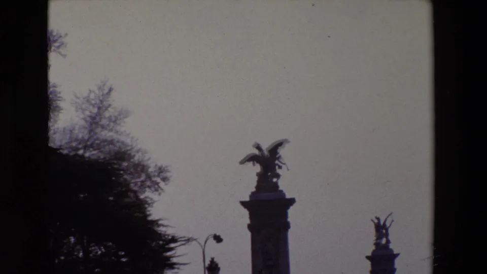 1969 Paris frame with a winged sculpture silhouetted on a high pedestal, tree branches at left, and another monument visible farther back.