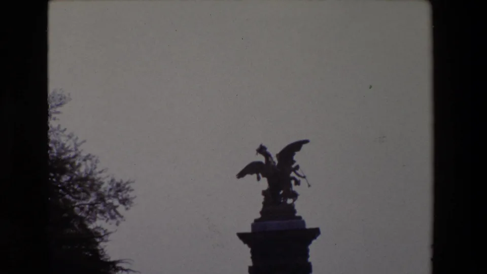 Closer 1969 Paris home-movie view of the winged sculpture silhouetted against the sky, with the pedestal top and tree branches visible at the left.