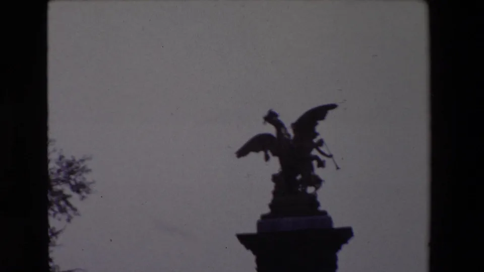 Paris 1969 8mm frame showing a dark silhouetted winged statue atop a column, with tree branches at the edge of the frame.