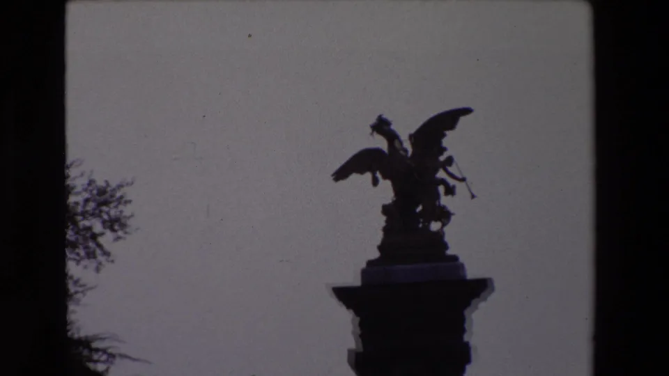 Paris 1969 home-movie frame with the winged statue still silhouetted on its column, framed by sky and partial trees.