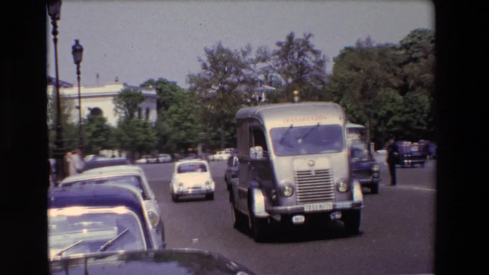 Paris 1969 street frame showing classic European automobiles, a light-colored truck with visible lettering, pedestrians, trees, and distant buildings.