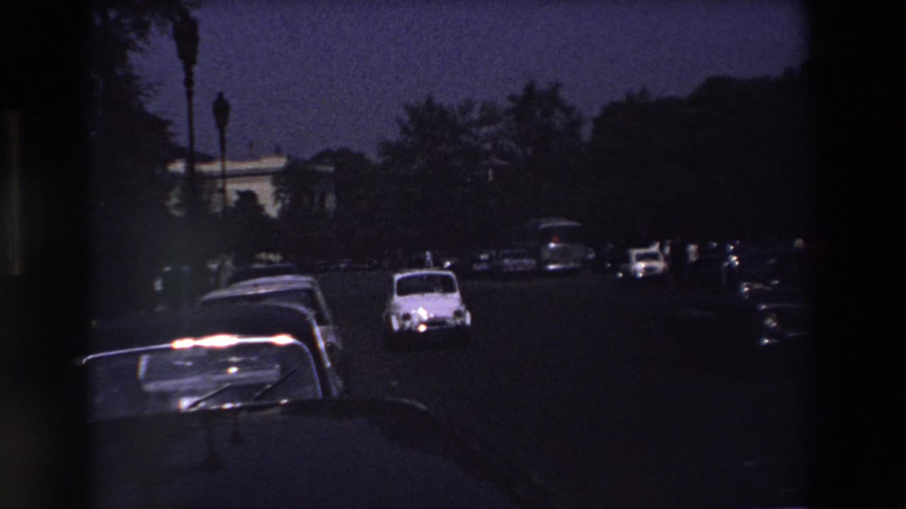 Dim 1969 Paris roadway with a white compact car approaching, dark parked cars along the curb, and trees framing the street.