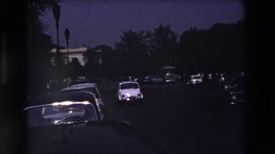 Dim 1969 Paris roadway with a white compact car approaching, dark parked cars along the curb, and trees framing the street.