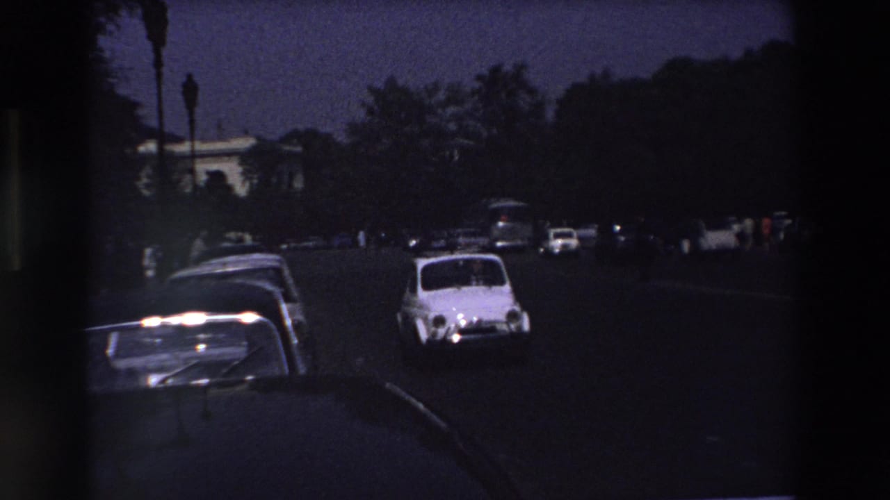 White compact car passing parked vehicles on a dark street.