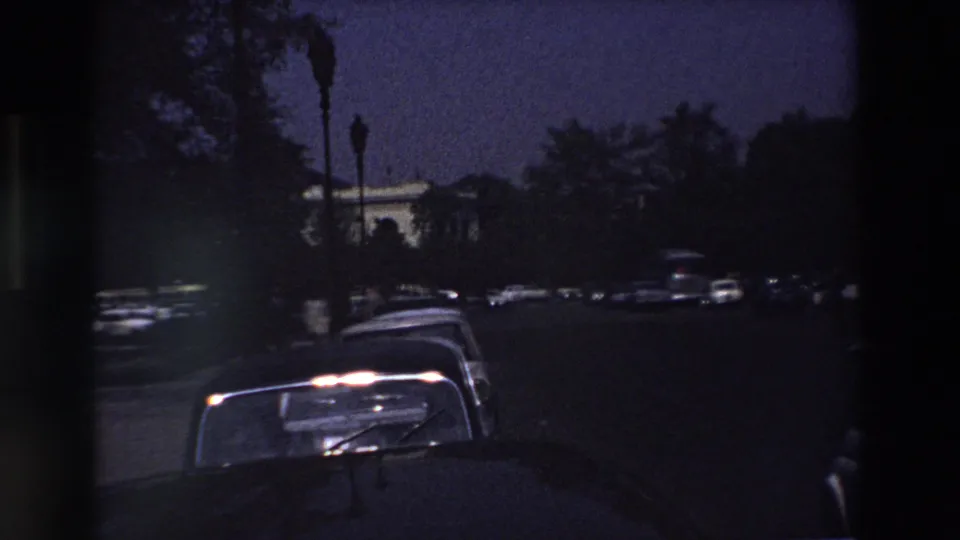 Late-dusk 1969 Paris street view with parked cars in the foreground, moving vehicles in the distance, and a pale building partly visible through trees.