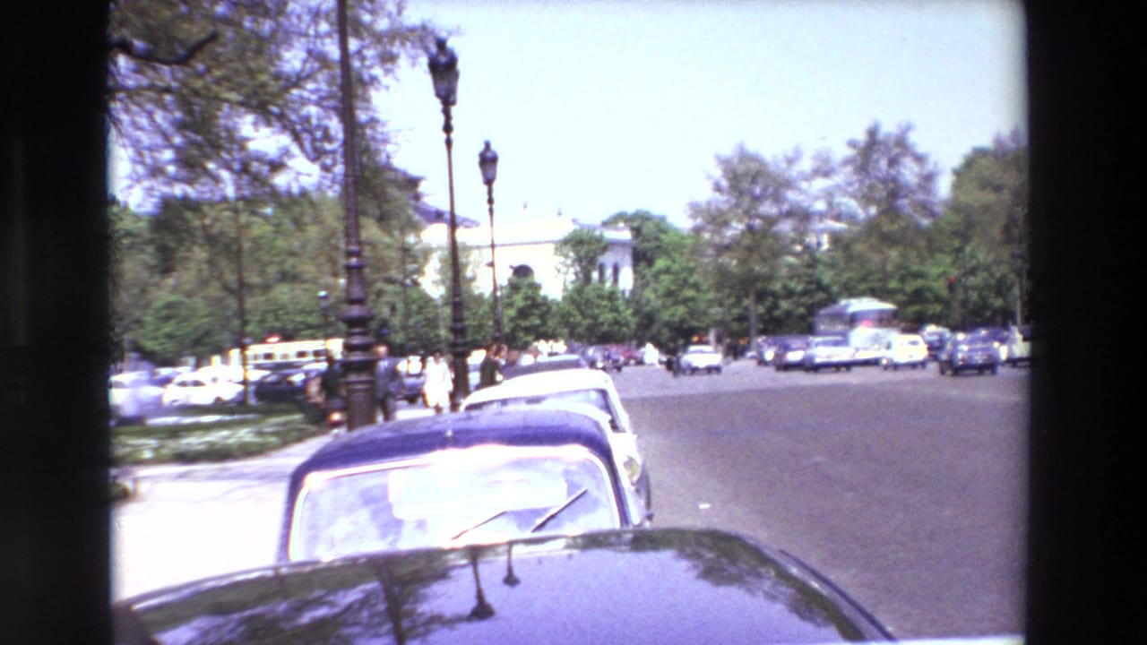 1969 Paris street view from a car, with a dark sedan foreground, parked cars along the curb, pedestrians near the crossing, trees, lamp posts, and distant traffic.