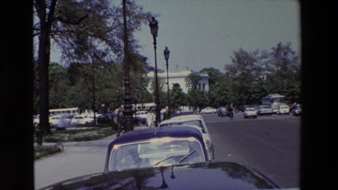 1969 Paris boulevard scene with parked cars close to the camera, pedestrians crossing near the curb, a white bus and several cars in traffic, and trees framing a pale building.