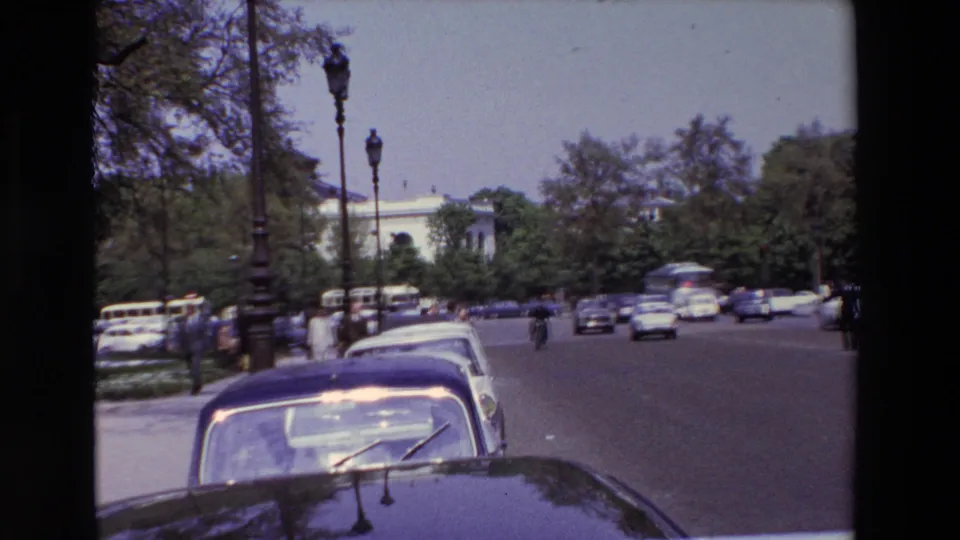 1969 Paris street view from a vehicle perspective, showing a dark car hood, a row of parked cars, pedestrians near the roadway, moving traffic, lamp posts, and leafy trees.