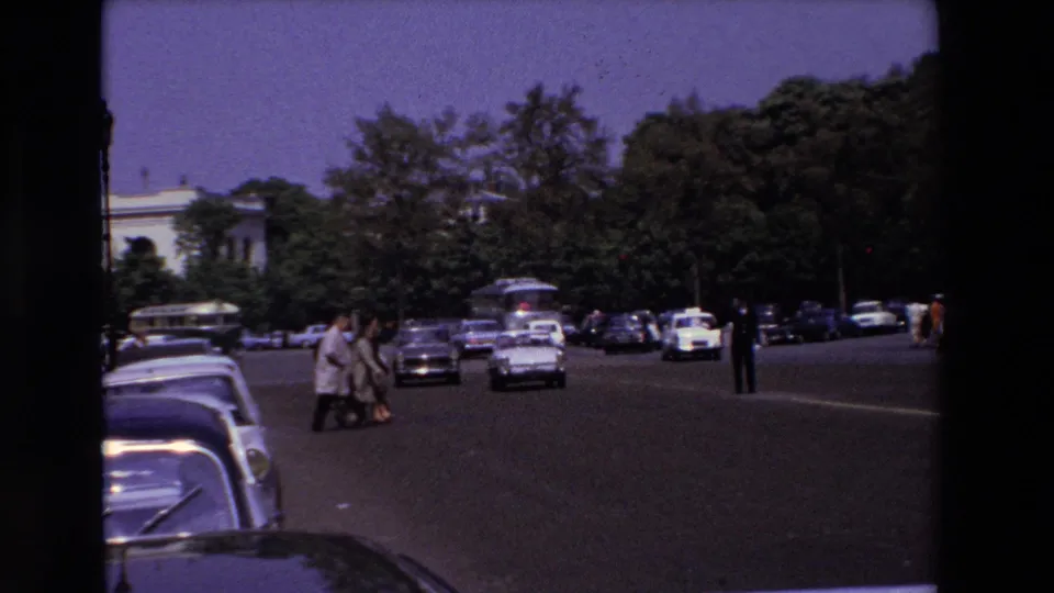 1969 Paris street frame with several pedestrians crossing in front of oncoming period cars, parked vehicles along the left curb, trees, lamp posts, and a pale building in the background.